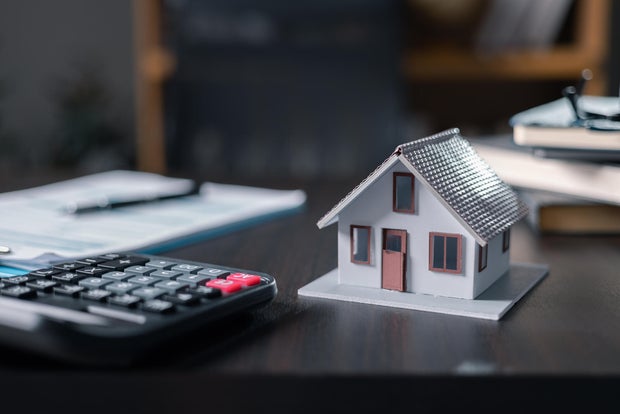 Model rumah, dokumen, dan uang tunai di desktop: real estat, pinjaman rumah, dan konsep investasi. Rumah kecil dengan kalkulator desain modern dan pena di latar belakang kantor untuk konsep pinjaman rumah 