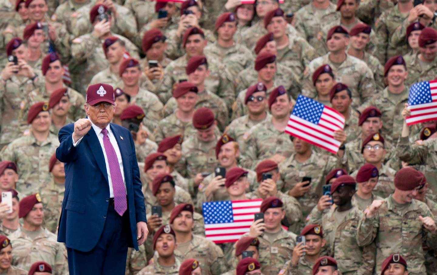 Presiden Donald Trump mengenakan topi bisbol dengan lencana militer dan mengangkat kepalan tangan. Dia menghadap jauh dari kerumunan pasukan di Fort Bragg yang mengenakan seragam bermotif kamuflase dan baret merah marun. Beberapa tentara mengangkat ponsel atau bendera Amerika mereka.