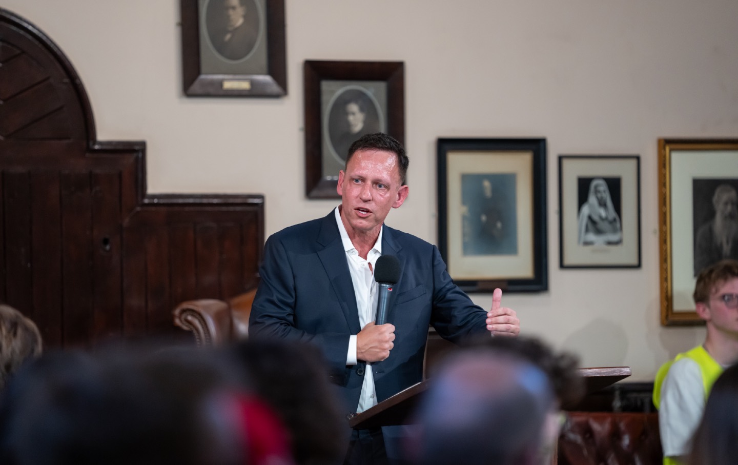 Peter Thiel berbicara di mimbar, memegang mikrofon di depan penonton di The Cambridge Union.